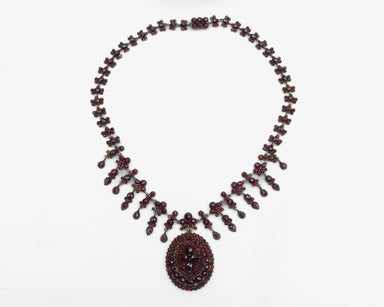 A vintage-style necklace featuring dark red faceted beads and a large circular pendant, laid flat on a white background.