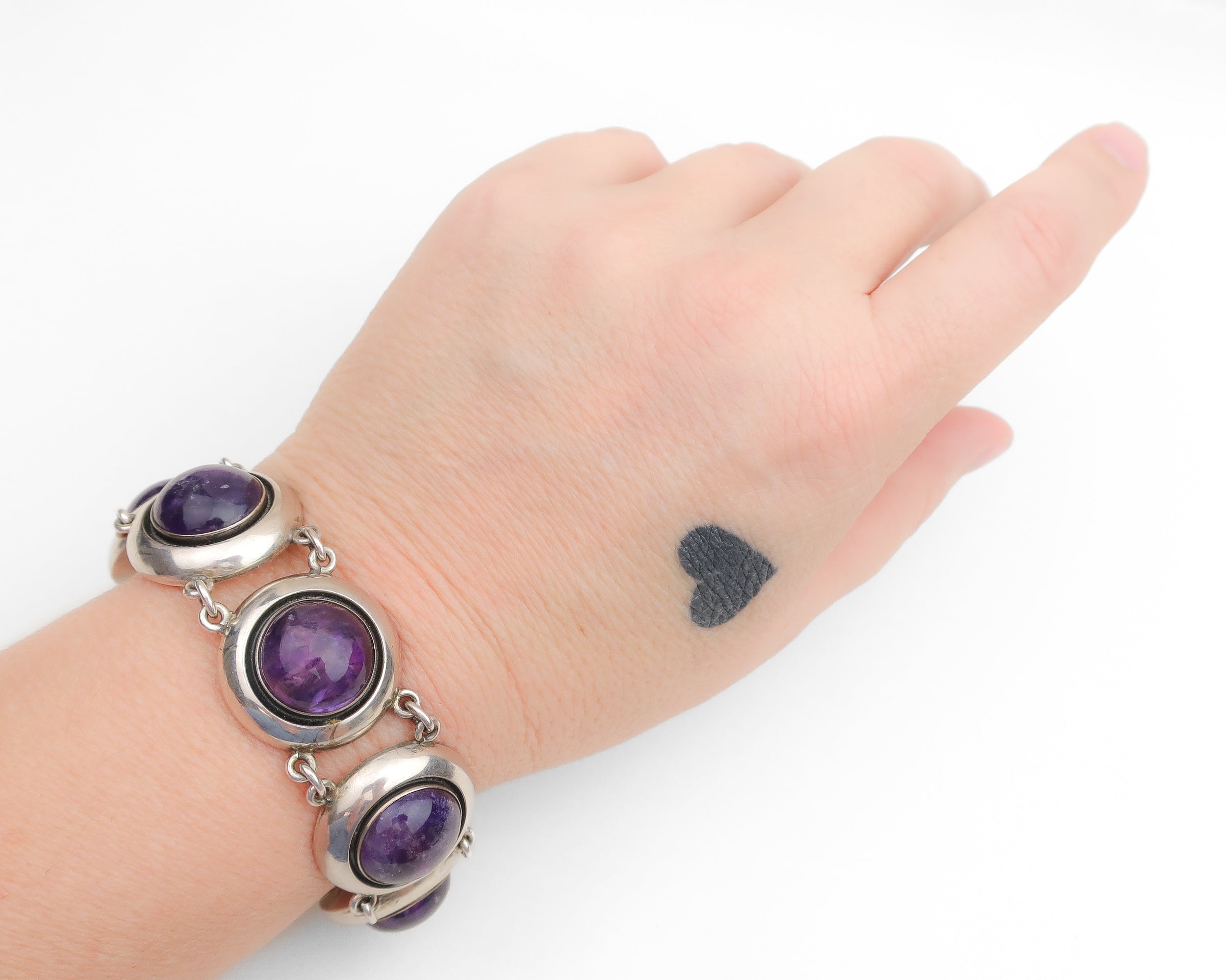 A close-up of a person's hand and wrist wearing a silver-toned bracelet with three oval purple amethyst stones, against a plain white background.
