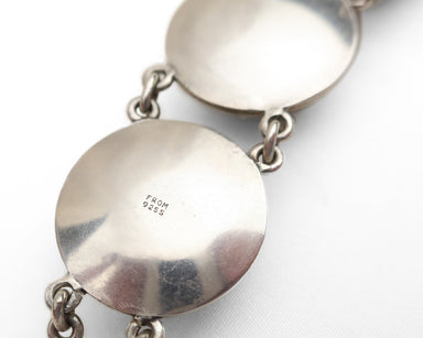 Close-up of a silver link bracelet showing a polished round disc link with a stamped 'FROM 925' hallmark on its flat surface, connected by small silver chains against a white background.